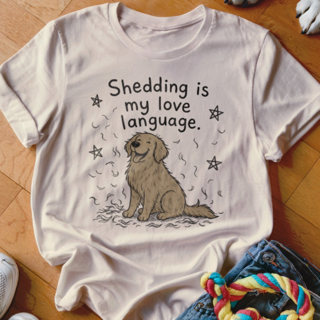 A cream-colored t-shirt with a graphic of a smiling dog surrounded by fur tufts and stars, reading Shedding is my love language.