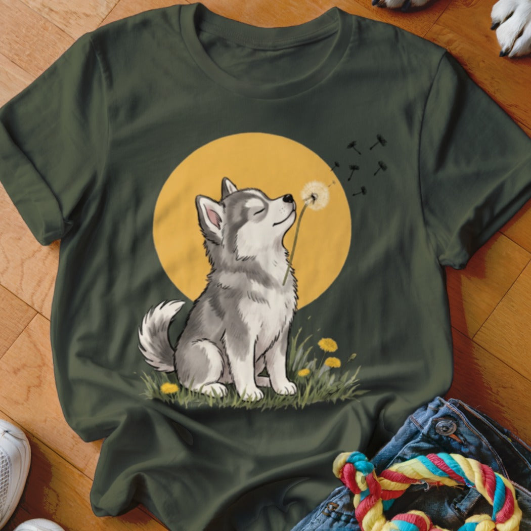 An olive green t-shirt featuring an illustration of a husky puppy blowing dandelion seeds against a yellow sun.