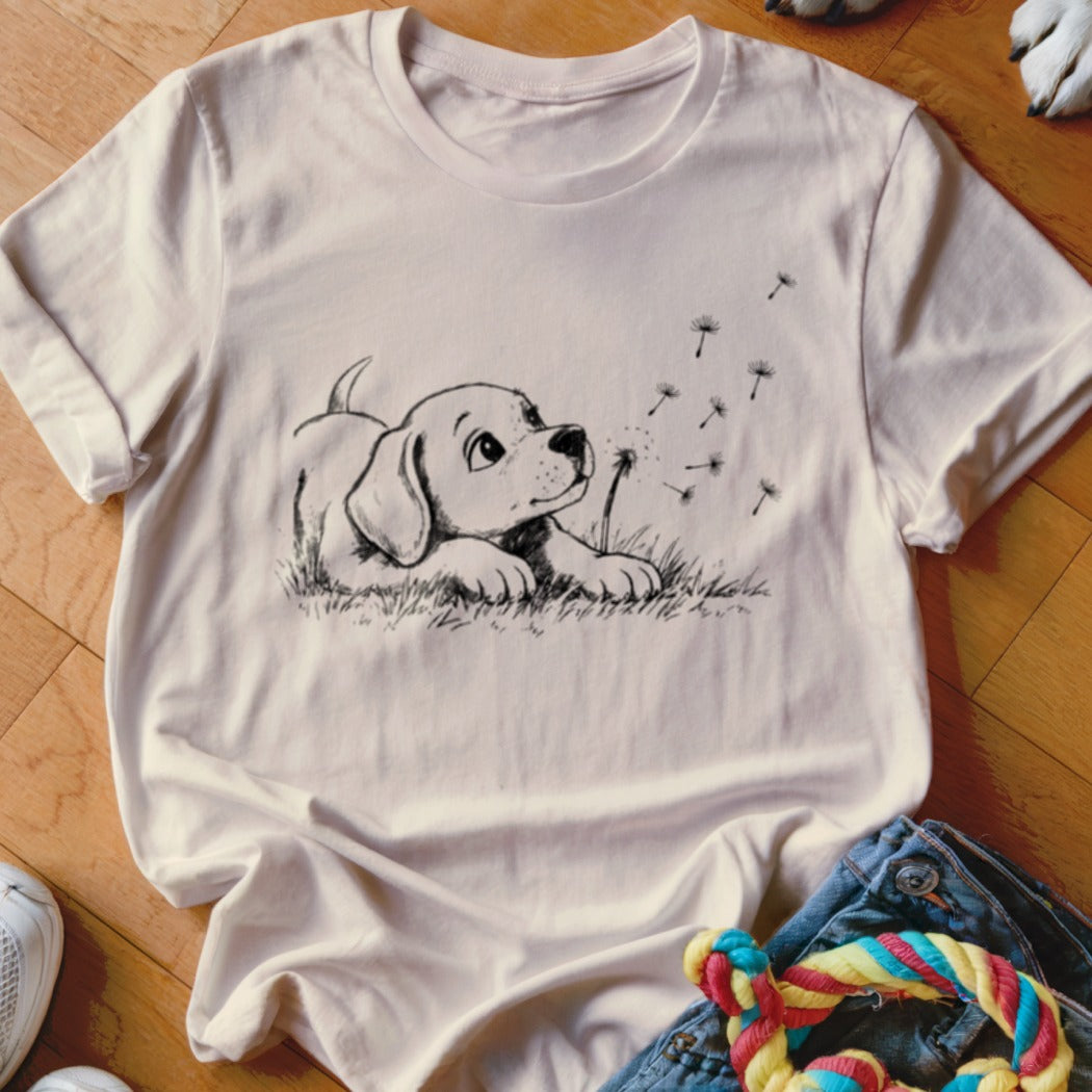 A light pink t-shirt features a black line drawing of a puppy watching floating dandelion seeds.
