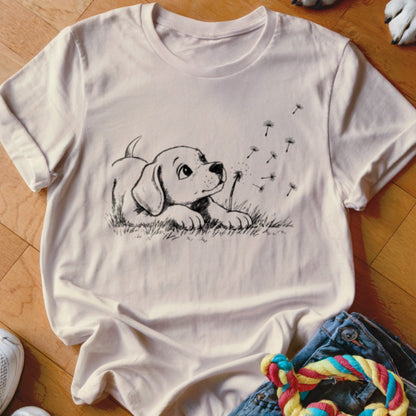 A light pink t-shirt features a black line drawing of a puppy watching floating dandelion seeds.