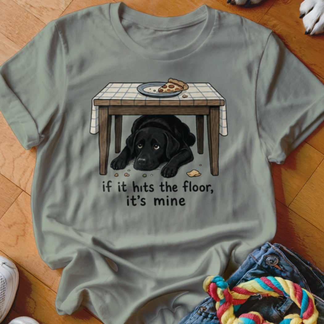 Black Labrador waits under a table for dropped pizza with text "if it hits the floor, it's mine".