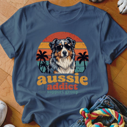 Dark blue t-shirt featuring a retro graphic of an Australian Shepherd wearing sunglasses before a sunset and palm trees, with the text Aussie Addict Support Group.