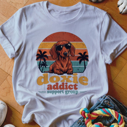 White t-shirt featuring a retro graphic of a dachshund in sunglasses with palm trees and the text doxie addict support group