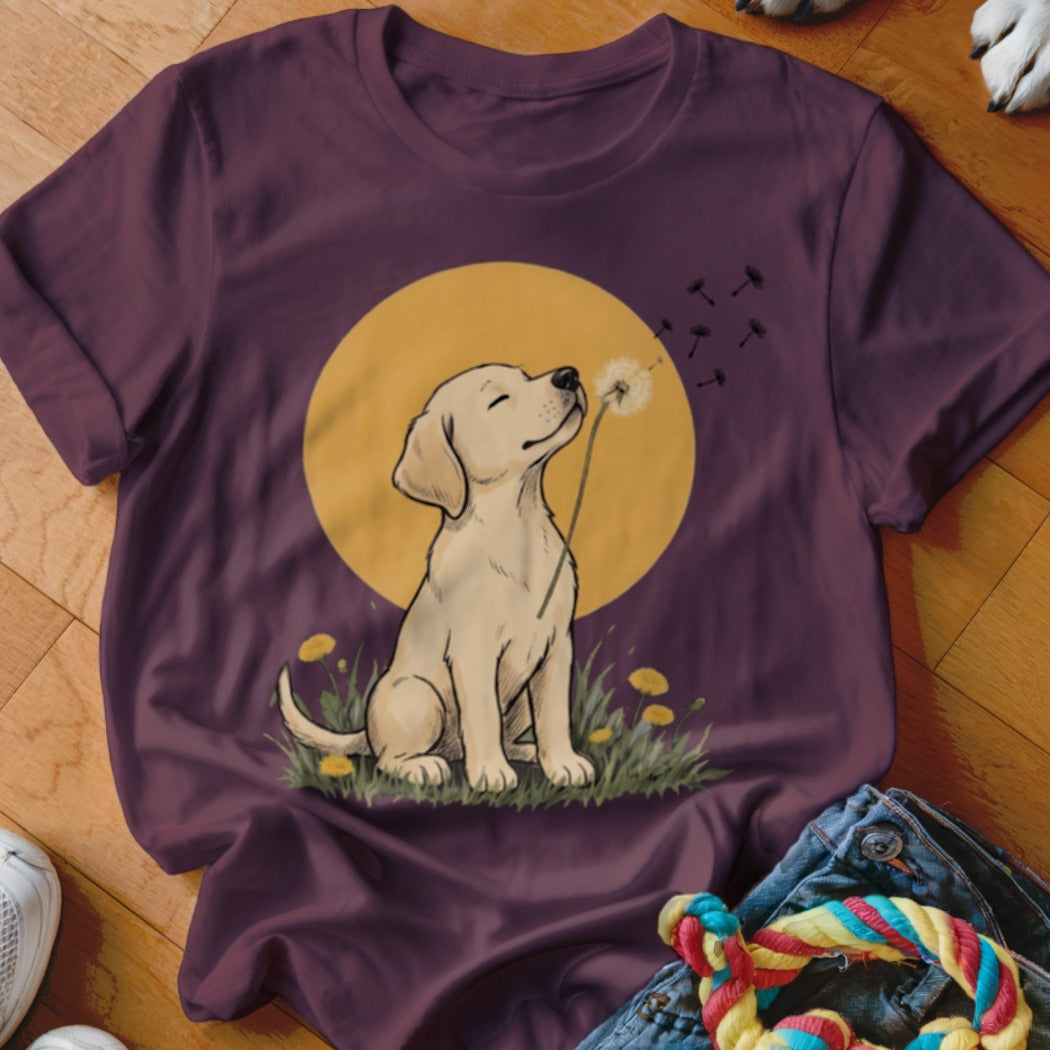 A dark purple short-sleeved t-shirt featuring an illustration of a yellow Labrador puppy blowing dandelion seeds in front of a yellow moon.