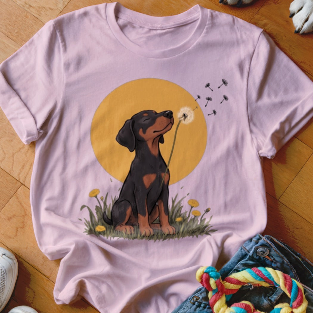 Light pink t-shirt featuring a cartoon Doberman puppy blowing dandelion seeds against a yellow sun.