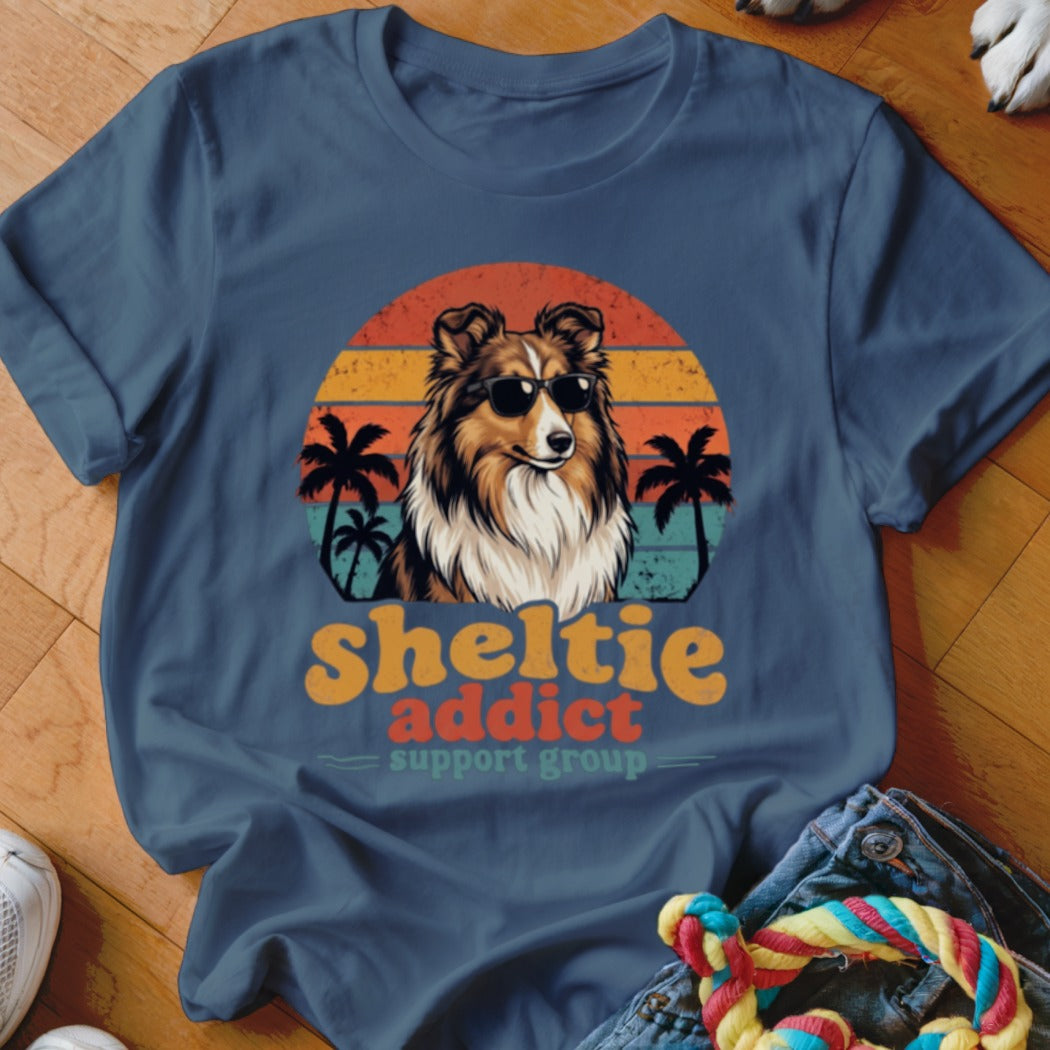 A blue crewneck t-shirt featuring a retro graphic of a Sheltie wearing sunglasses in front of a sunset and palm trees, with the text "sheltie addict support group."