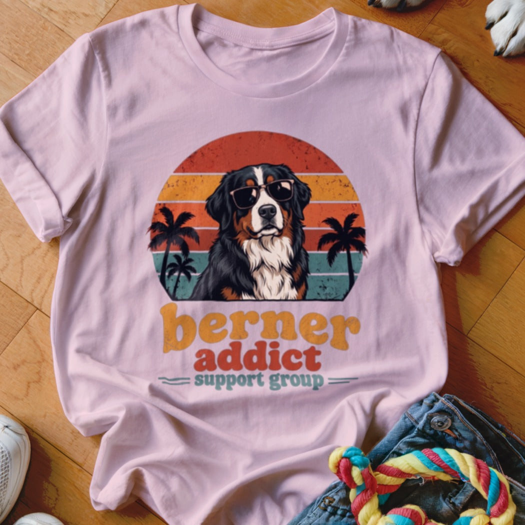 Light pink t-shirt featuring a Bernese Mountain Dog wearing sunglasses in front of a retro sunset and palm tree graphic with text reading berner addict support group.