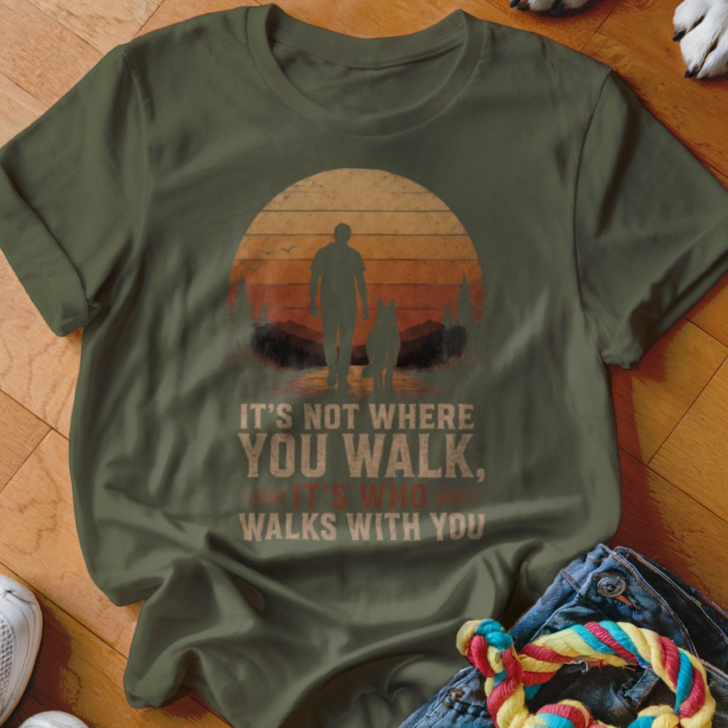 Olive t-shirt with a graphic of a person and dog walking toward a sunset. Text reads "It's not where you walk, it's who walks with you."