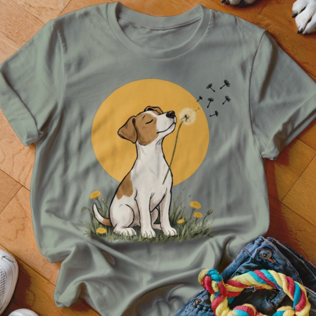 A sage green t-shirt featuring an illustration of a dog blowing dandelion seeds against a yellow sun.
