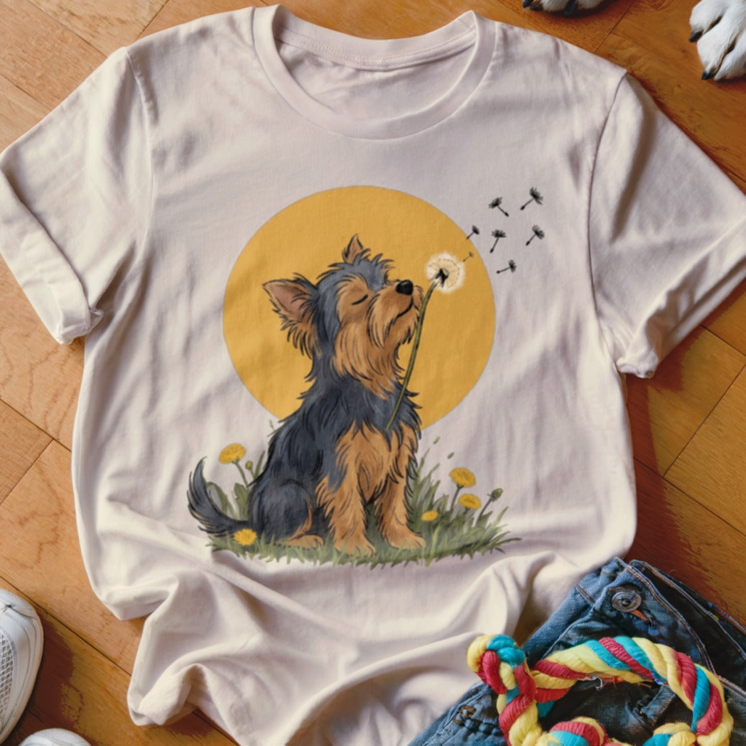 A light cream t-shirt features a graphic of a Yorkshire terrier blowing dandelion seeds against a yellow circle.