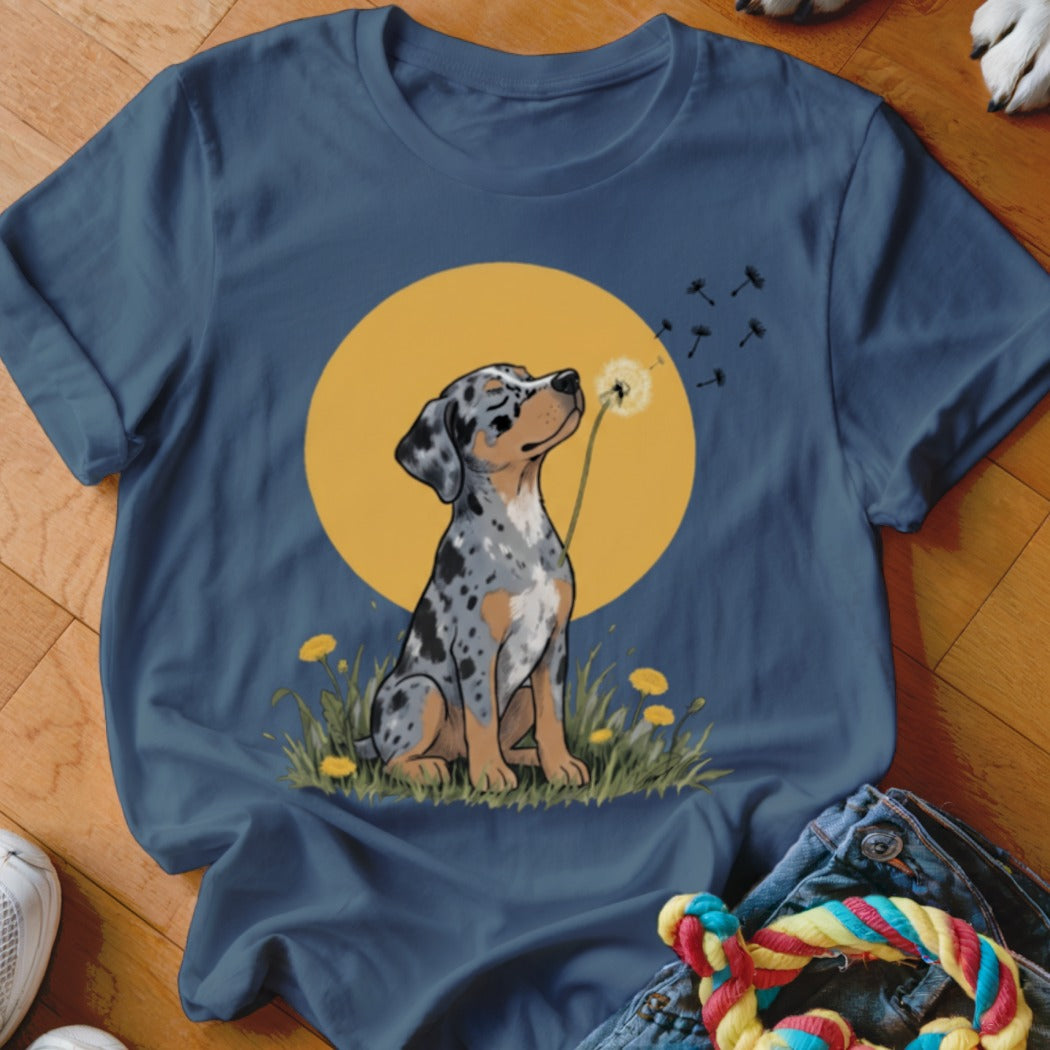 A dark blue t-shirt featuring a printed illustration of a merle puppy blowing a dandelion against a large yellow circle.