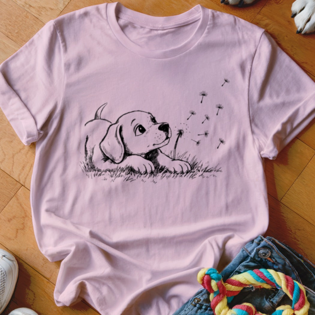 A pink t-shirt with a black line drawing of a puppy watching floating dandelion seeds.
