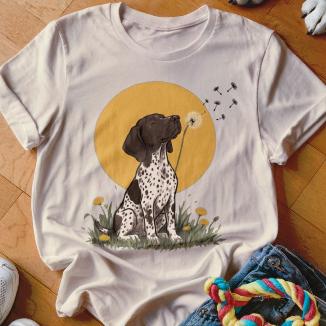 A cream-colored t-shirt featuring an illustration of a spotted dog blowing a dandelion against a yellow circle background.