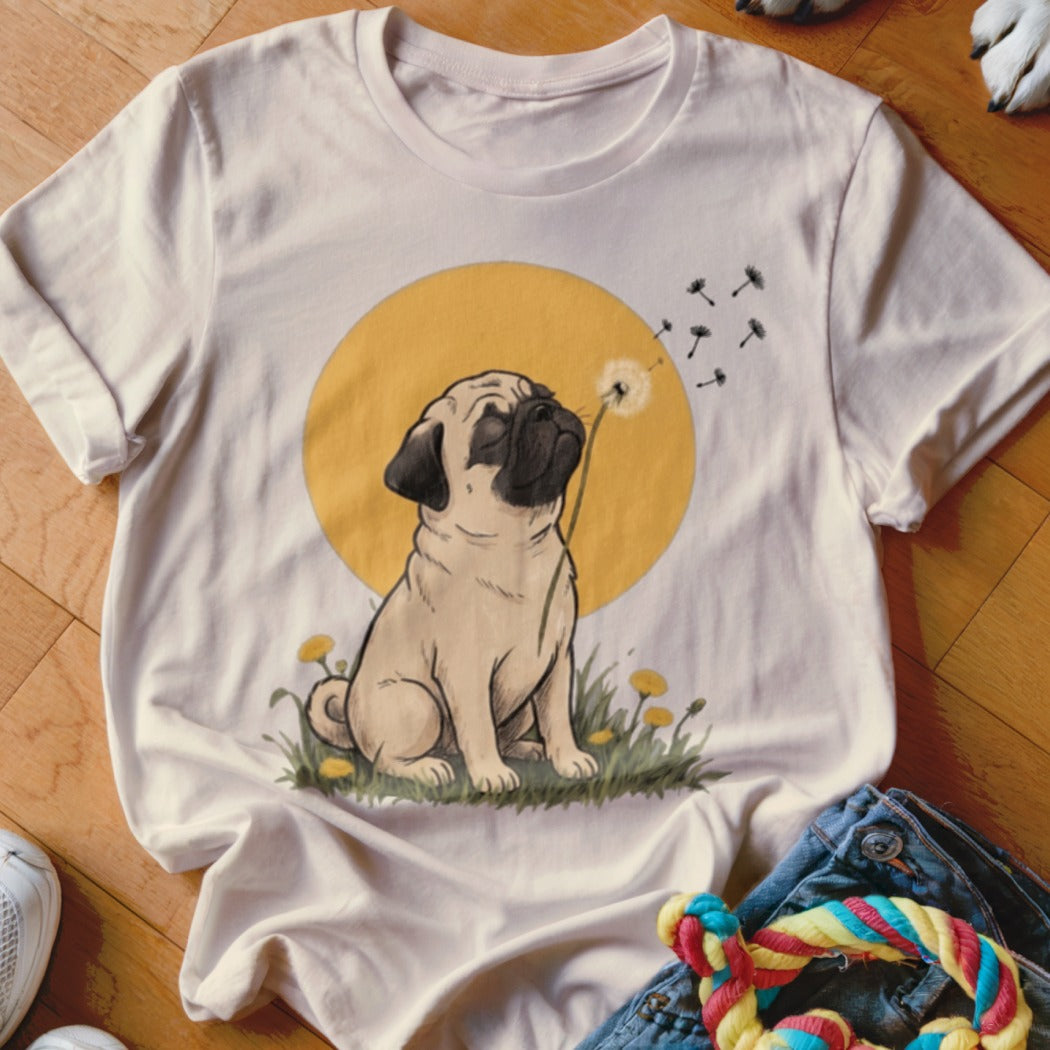 A light cream t-shirt features a graphic of a pug blowing dandelion seeds against a yellow circle background.