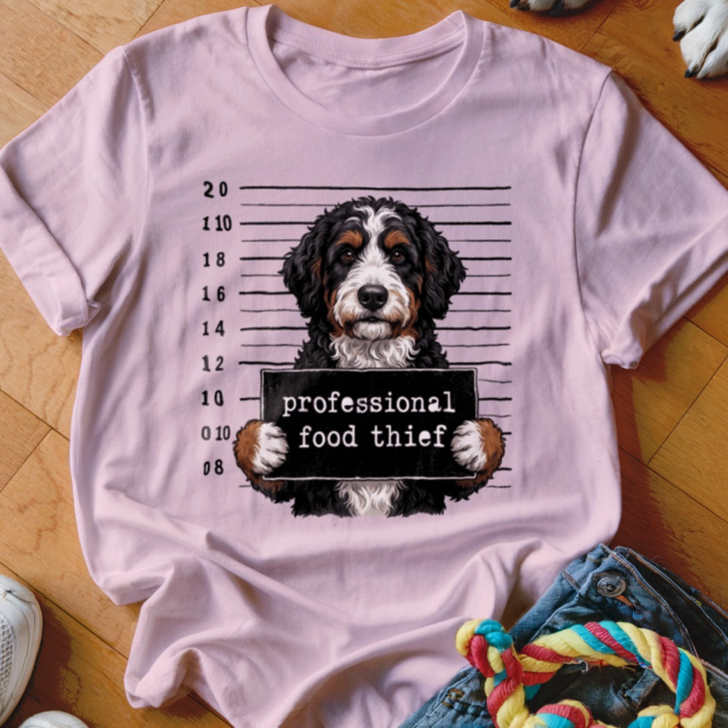 Bernese Mountain Dog mugshot with "professional food thief" sign on pink t-shirt.