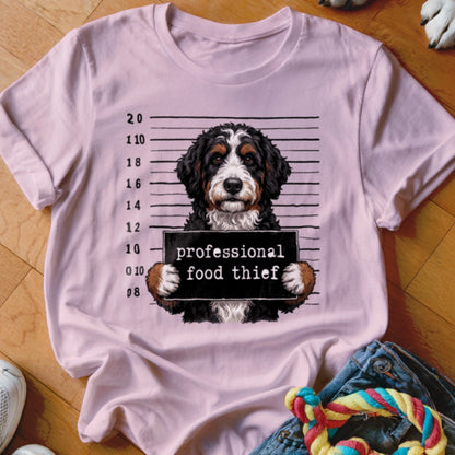 Bernese Mountain Dog mugshot with "professional food thief" sign on pink t-shirt.
