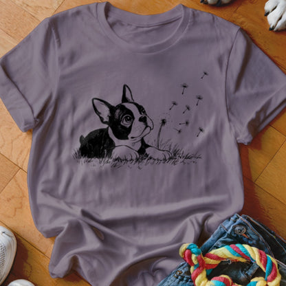 A purple crewneck t-shirt featuring a black-and-white graphic of a Boston Terrier in grass blowing dandelion seeds.