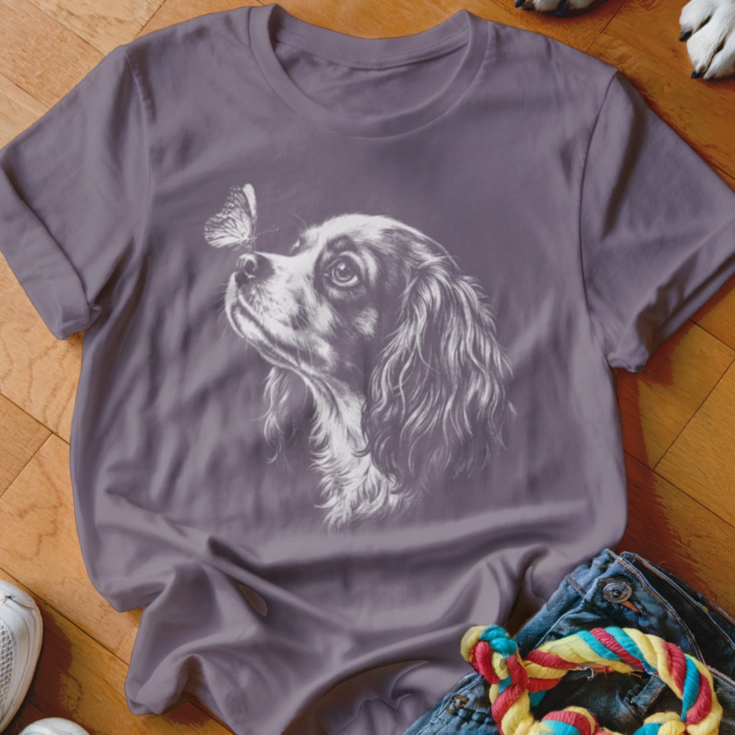 A purple t-shirt featuring a white sketch graphic of a King Charles Spaniel looking at a butterfly on its nose.