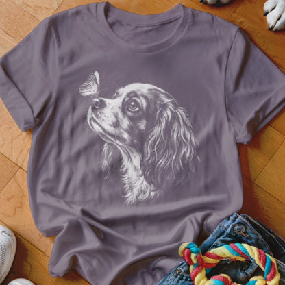 A purple t-shirt featuring a white sketch graphic of a King Charles Spaniel looking at a butterfly on its nose.