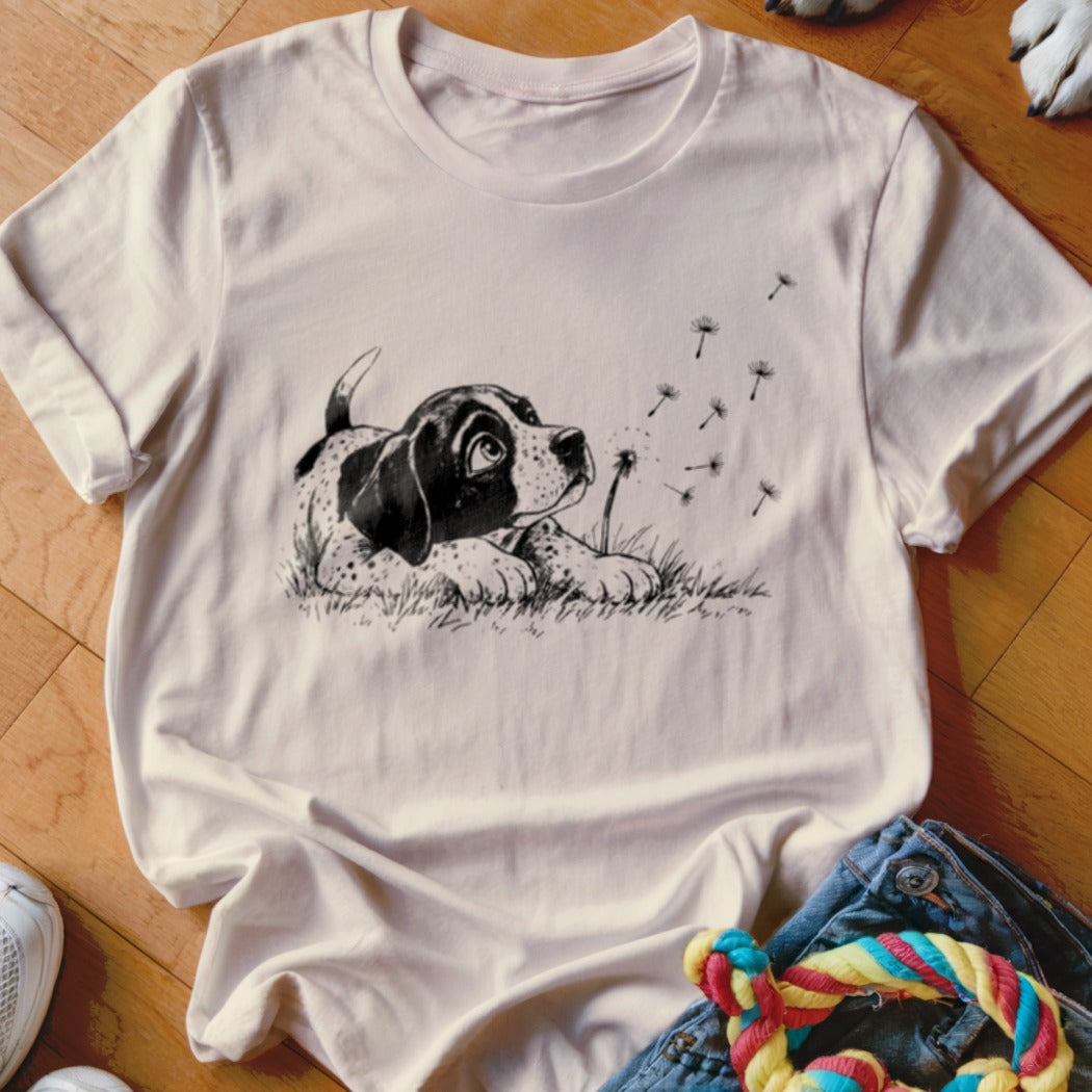 A light pink crewneck t-shirt featuring a black and white sketch of a puppy watching dandelion seeds blow away.
