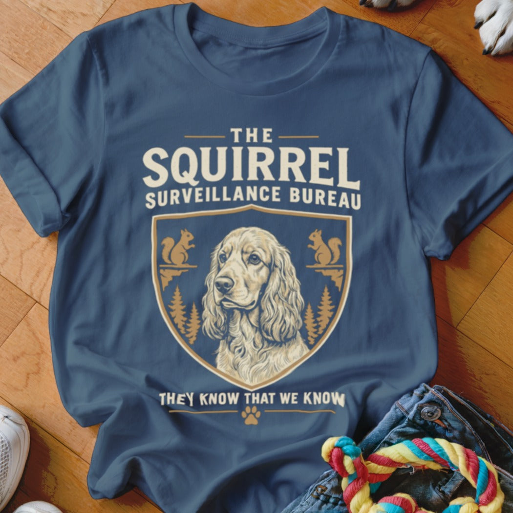 A blue t-shirt printed with a crest featuring a spaniel, squirrels, trees, and text reading "The Squirrel Surveillance Bureau."