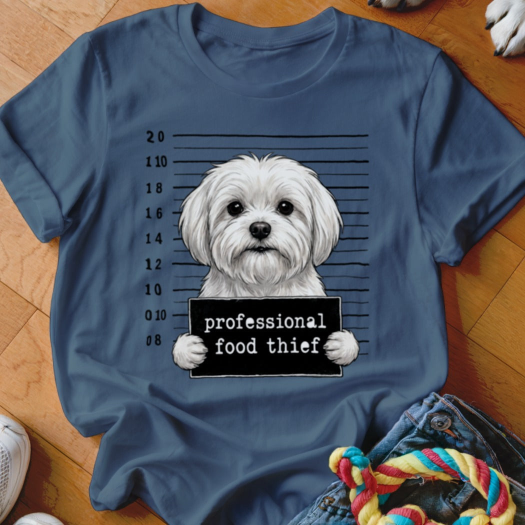 Maltese dog mugshot holding "professional food thief" sign on blue t-shirt.