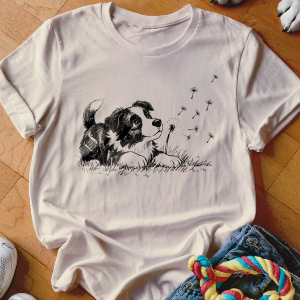 A cream-colored t-shirt with a black line art graphic of a puppy looking at dandelion seeds.