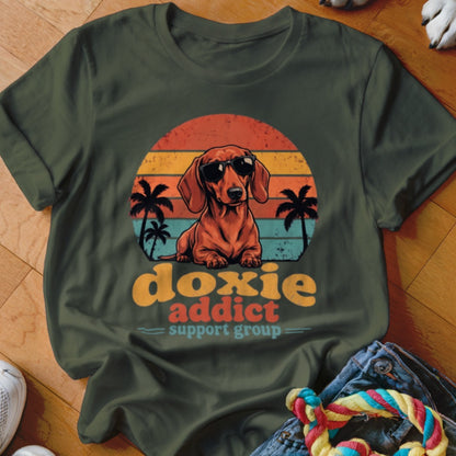 Olive green t-shirt featuring a retro graphic of a dachshund in sunglasses against a sunset, with palm trees and the text Doxie Addict Support Group.