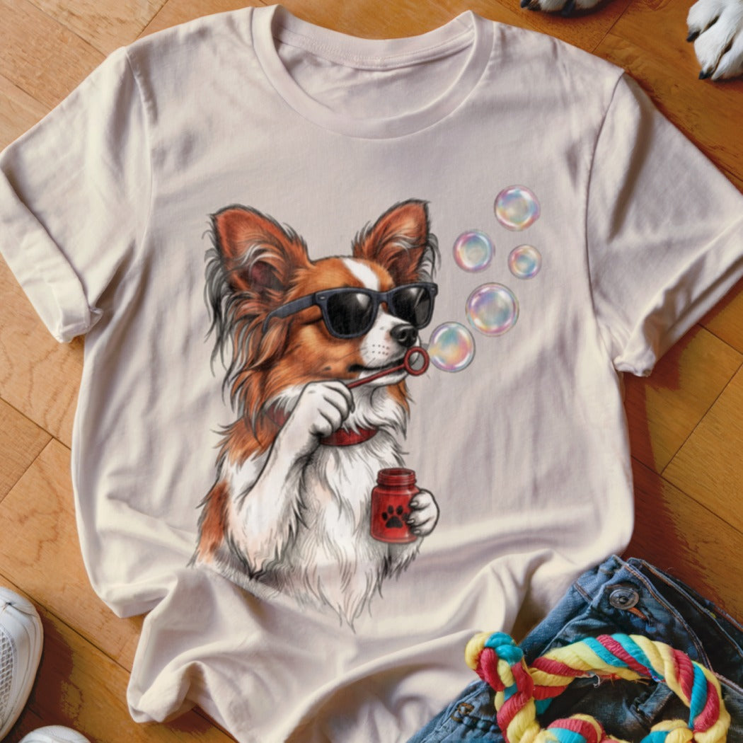 A light beige t-shirt featuring an illustration of a Papillon dog wearing sunglasses and blowing bubbles.