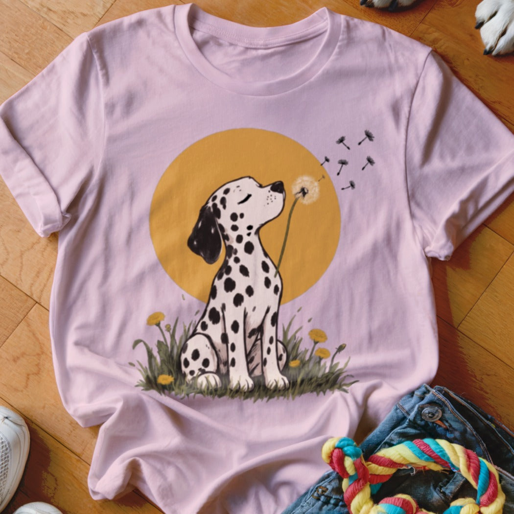 Light pink t-shirt featuring an illustration of a dalmatian blowing dandelion seeds in a grassy field.