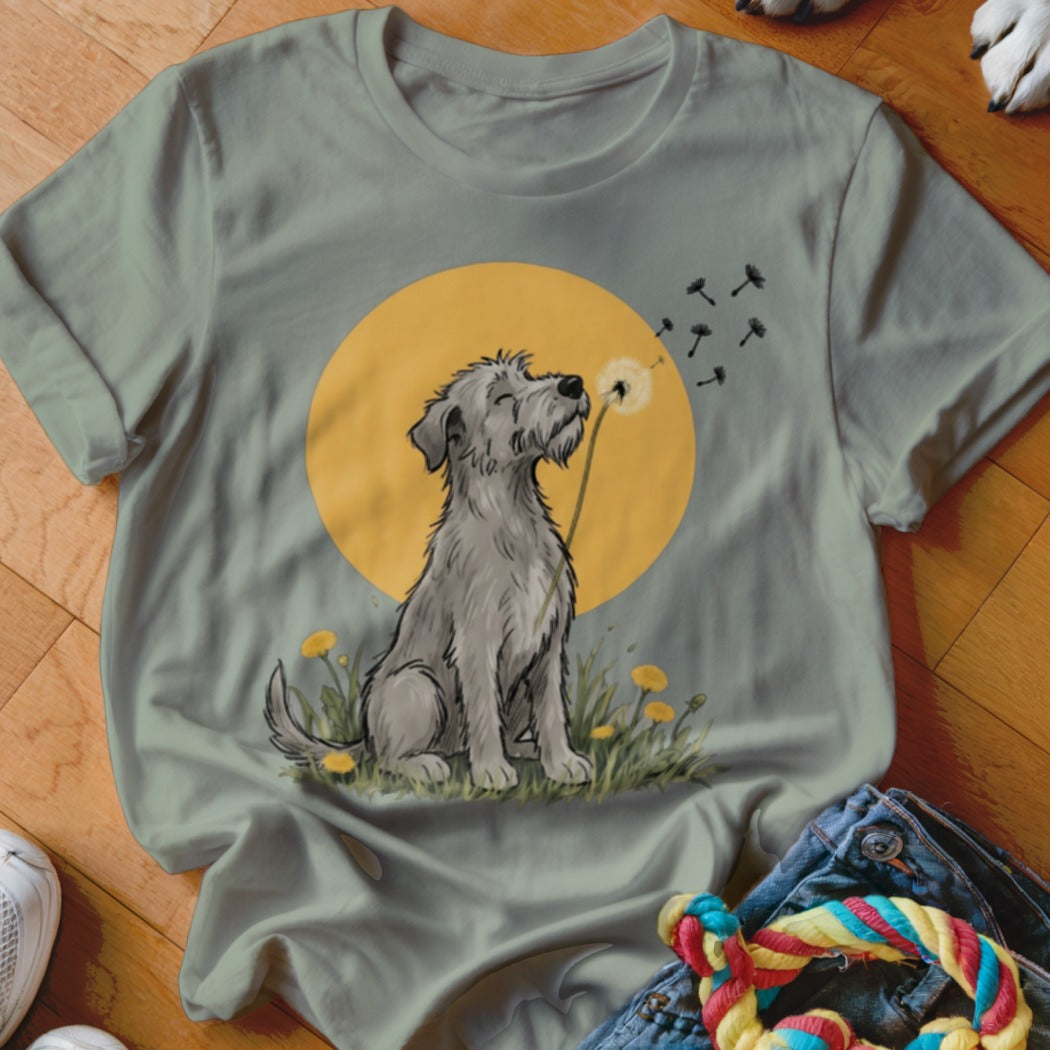 Sage green t-shirt featuring a cartoon illustration of a grey dog blowing dandelion seeds against a yellow sun.