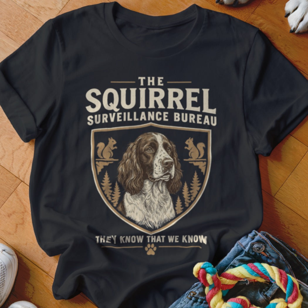 A black crewneck t-shirt featuring a graphic of a dog inside a crest with squirrel illustrations and text reading THE SQUIRREL SURVEILLANCE BUREAU.