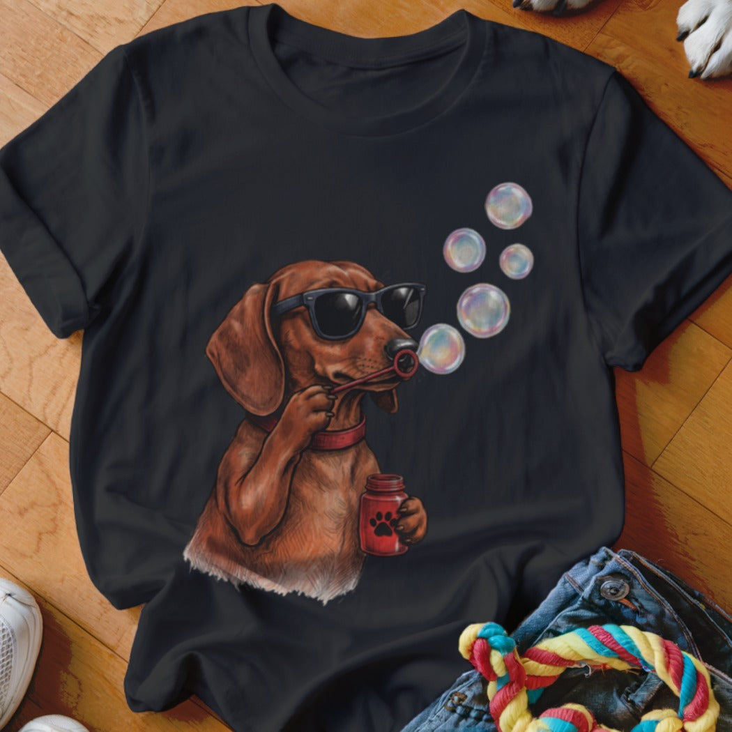 A black t-shirt featuring a graphic of a brown dachshund wearing sunglasses and blowing soap bubbles.