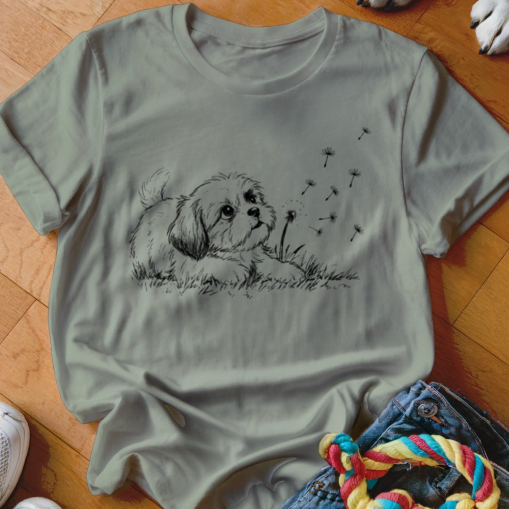 A sage green t-shirt featuring a black line drawing of a puppy watching dandelion seeds blow away.