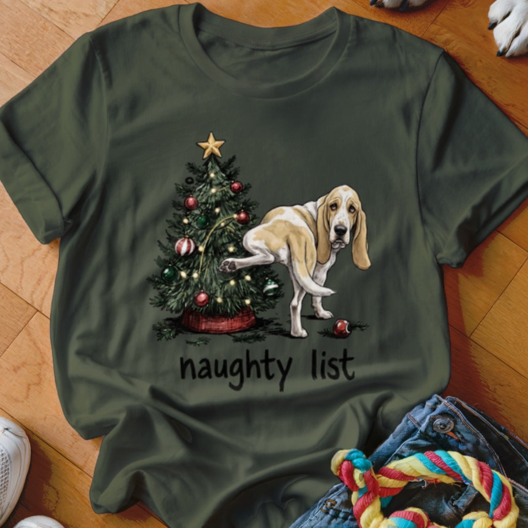 Basset hound near Christmas tree with text "naughty list" on dark green shirt.