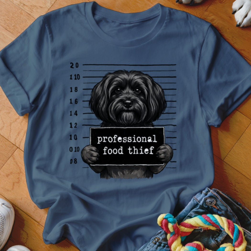 Black dog in mugshot holding "professional food thief" sign on a navy t-shirt.