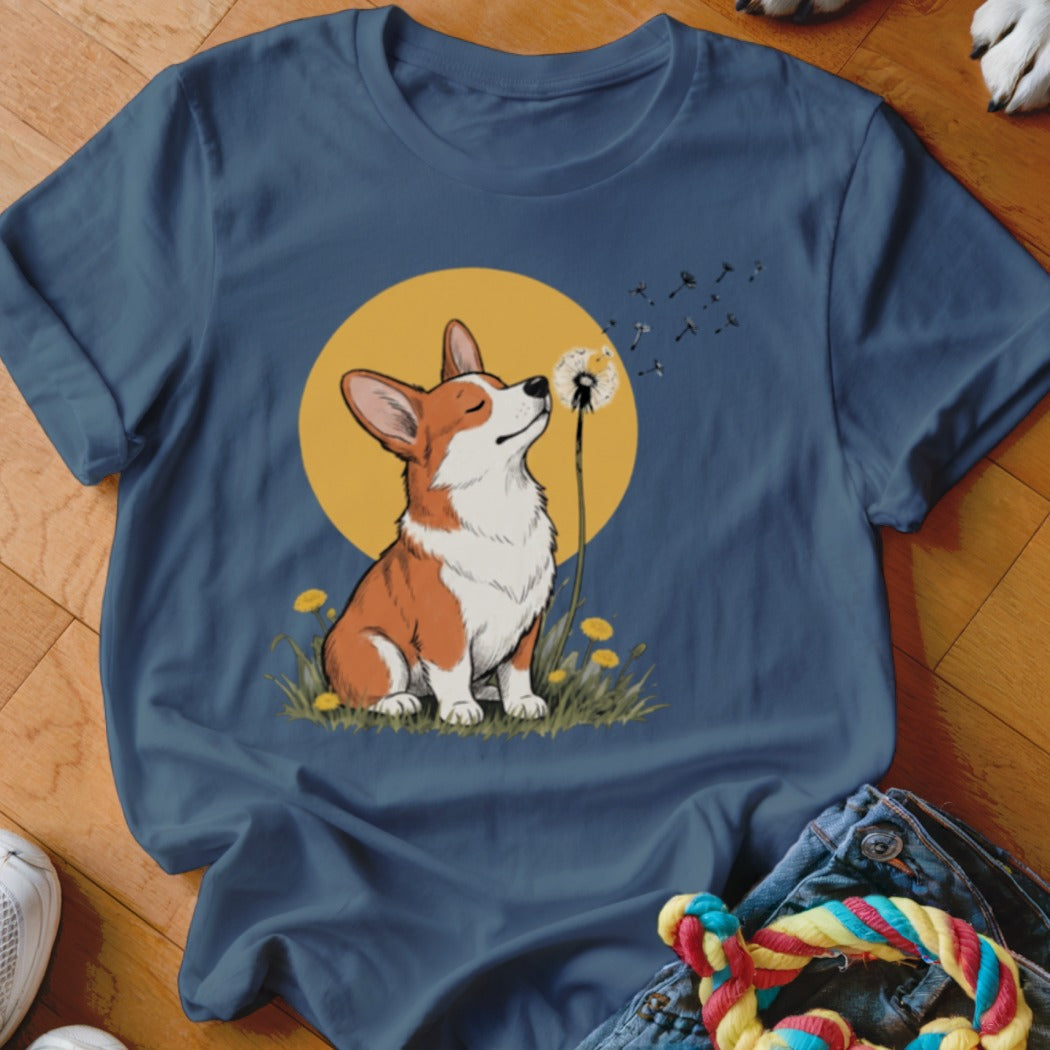 A blue t-shirt printed with a graphic of a corgi blowing dandelion seeds.