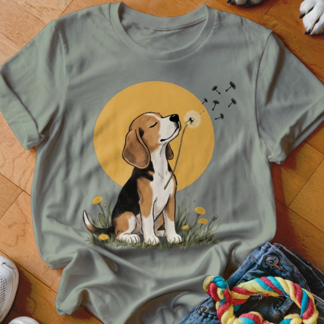 A sage green t-shirt features a graphic of a beagle blowing dandelion seeds against a yellow circle background.