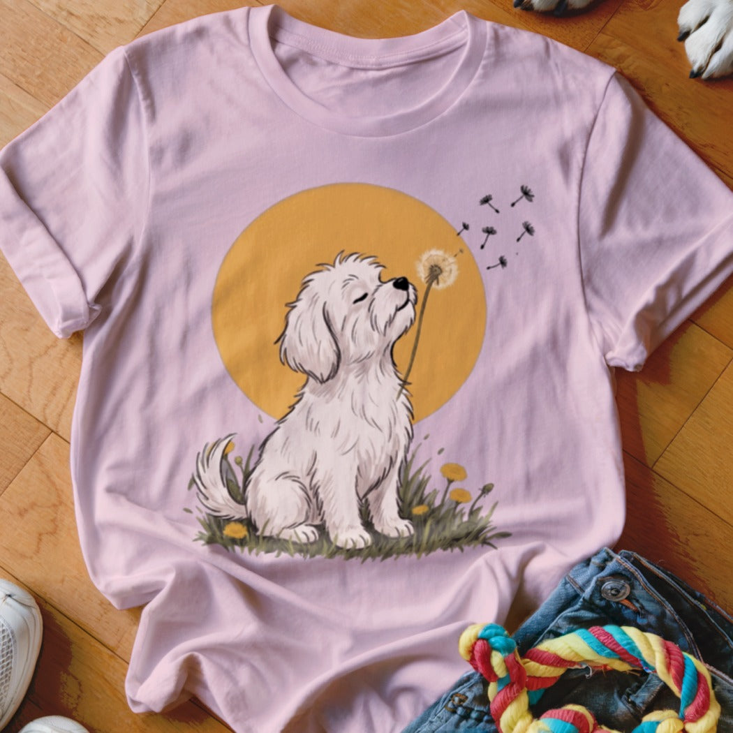 A light pink t-shirt features a graphic of a white fluffy dog blowing dandelion seeds against a yellow circle.