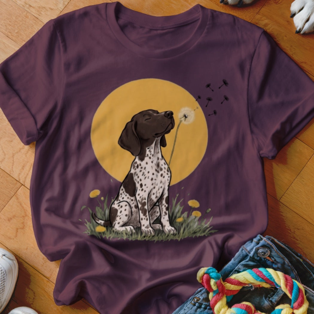 Maroon t-shirt featuring an illustration of a spotted puppy sitting in the grass blowing dandelion seeds.