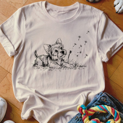 A light cream t-shirt printed with a black ink drawing of a small terrier puppy looking at dandelion seeds.