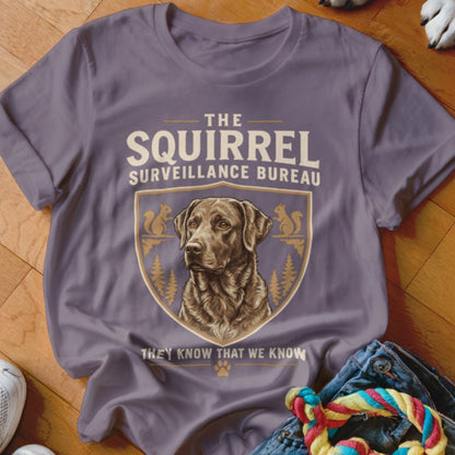 A purple crewneck t-shirt featuring a retro graphic of a dog, squirrels, and the text The Squirrel Surveillance Bureau.