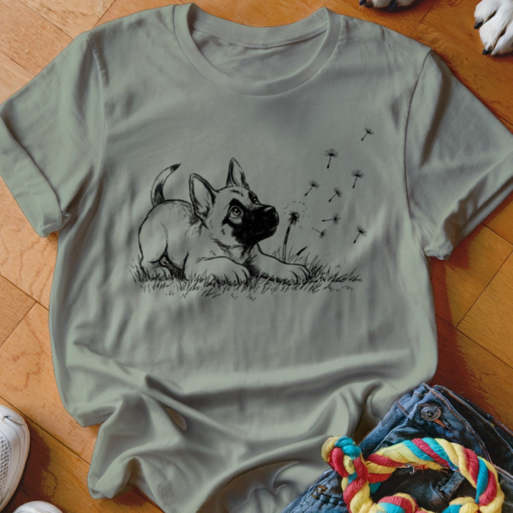 An olive green crewneck t-shirt featuring a black graphic of a puppy watching dandelion seeds blow in the wind.