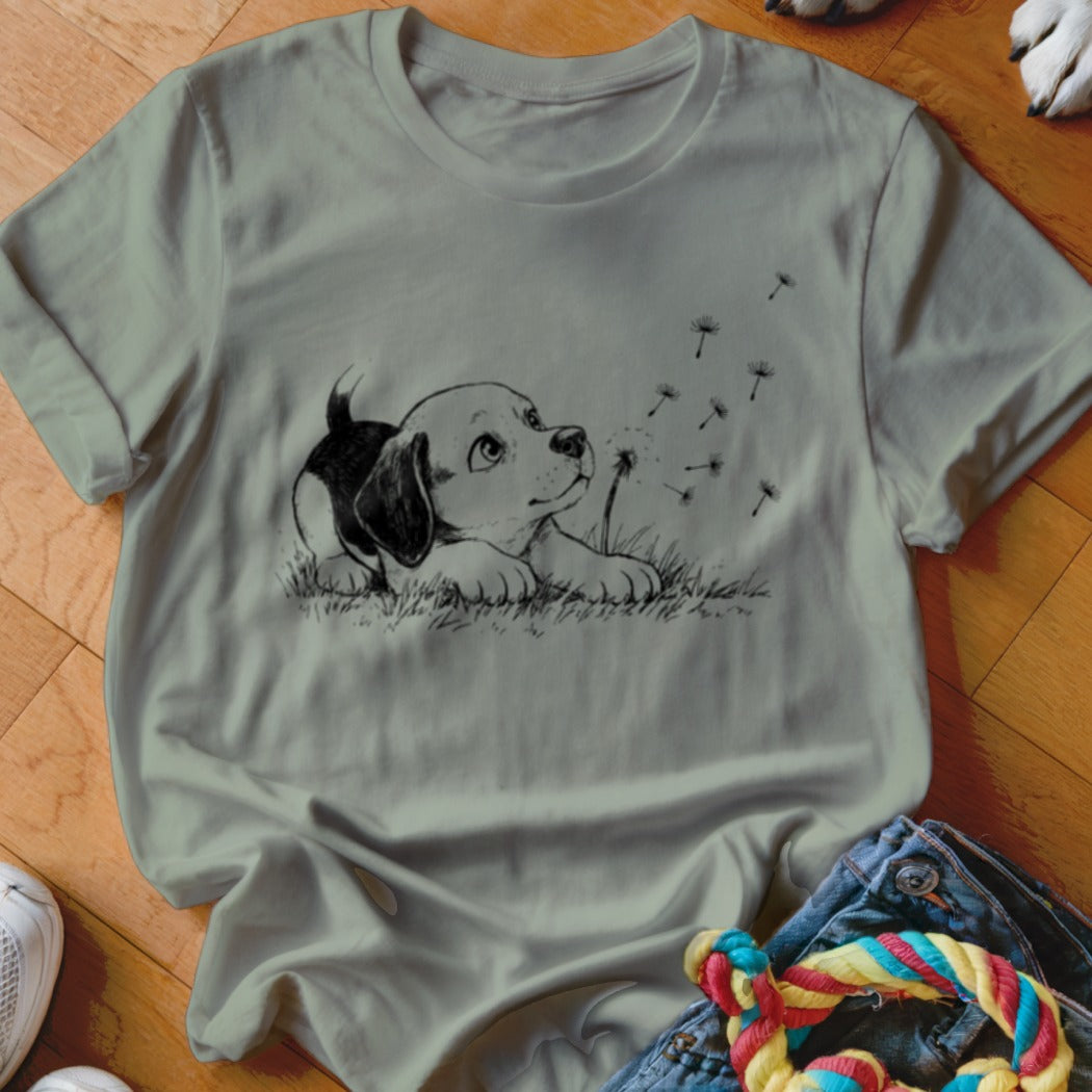 Sage green crewneck t-shirt featuring a black-and-white sketch of a puppy watching dandelion seeds blow away.