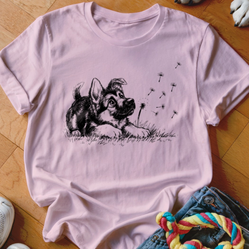 A pink t-shirt featuring a black and white sketch of a puppy watching dandelion seeds blow away.