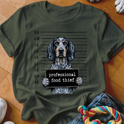 Dog with mugshot sign reading "professional food thief" on green t-shirt.