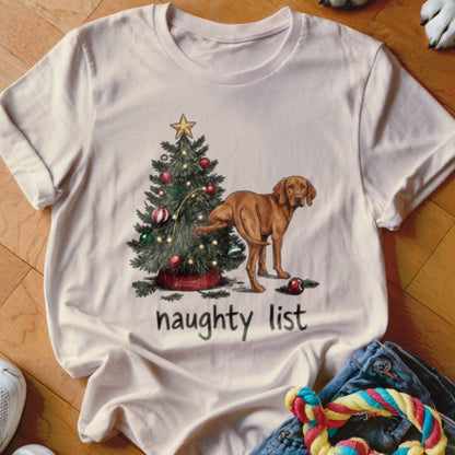 Light-colored t-shirt featuring a graphic of a brown dog urinating on a Christmas tree with text that says naughty list.
