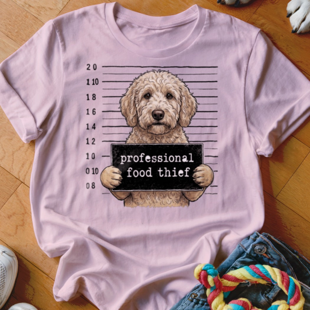 Goldendoodle mugshot with "professional food thief" sign on a light pink t-shirt.