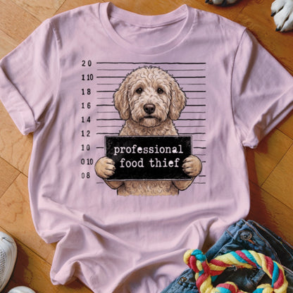 Goldendoodle mugshot with "professional food thief" sign on a light pink t-shirt.