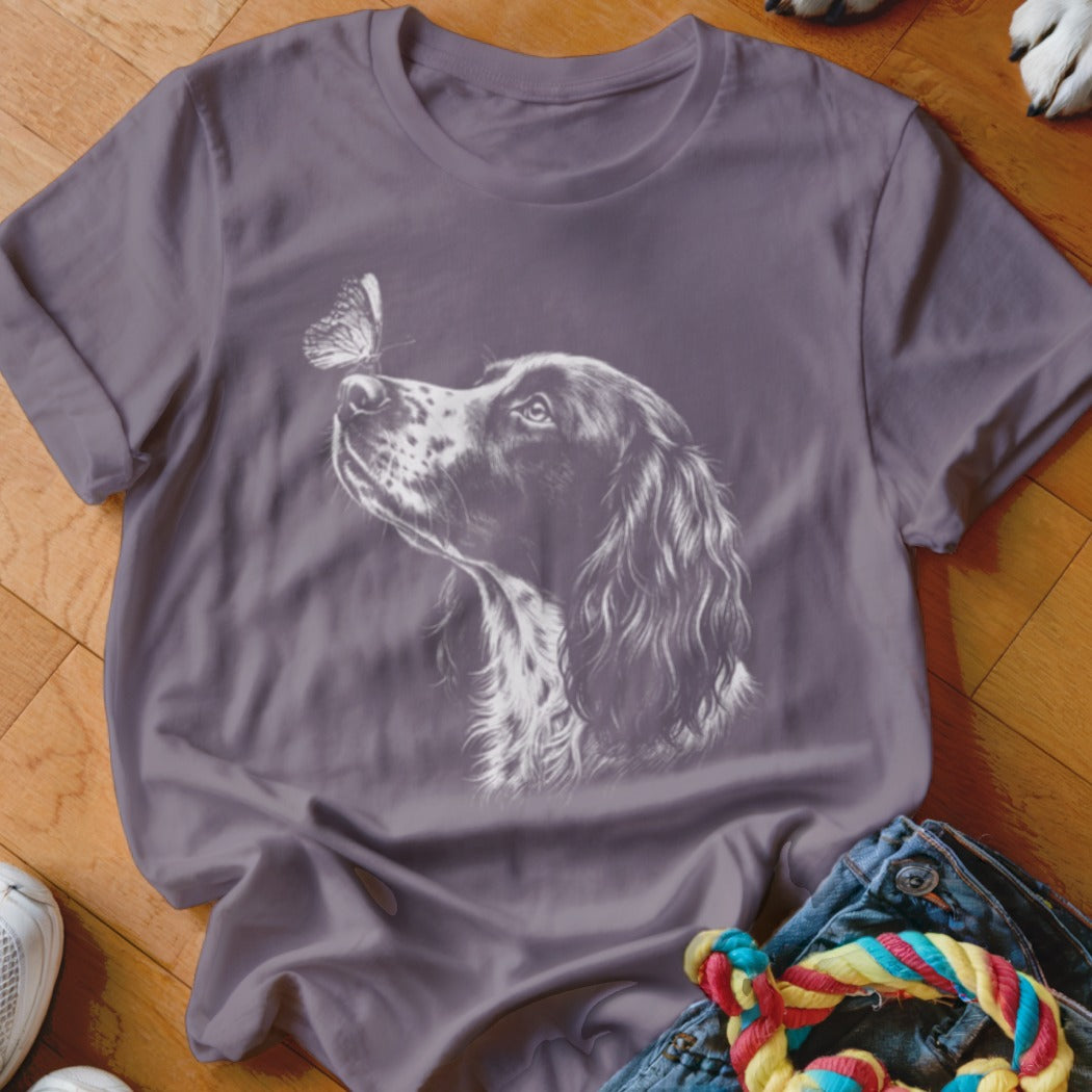 A purple crewneck t-shirt featuring a white line drawing of a spaniel looking at a butterfly on its nose.